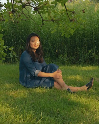 Stephanie smiling and sitting under a green canopy of trees on a patch of grass in a blue denim dress. 